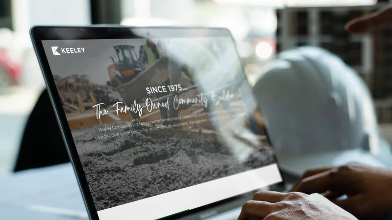 Laptop displaying the Keeley Companies website on a construction jobsite with a hard hat in the background.