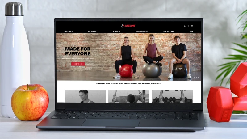 Laptop displaying the Lifeline Fitness website homepage featuring home workout equipment, with fitness accessories including dumbbells, a water bottle, and an apple arranged around the computer to represent an active, healthy lifestyle.