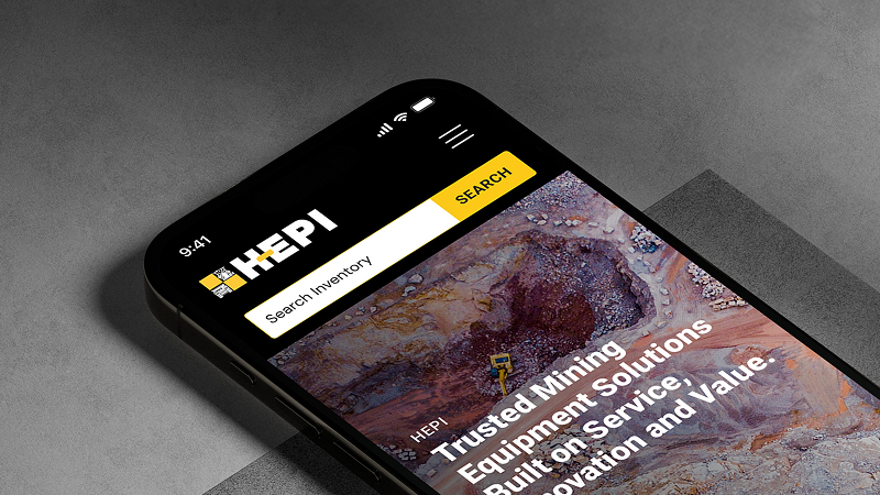 Close-up of a smartphone showing the HEPI website, featuring a search bar and an overhead image of a mining site with equipment.