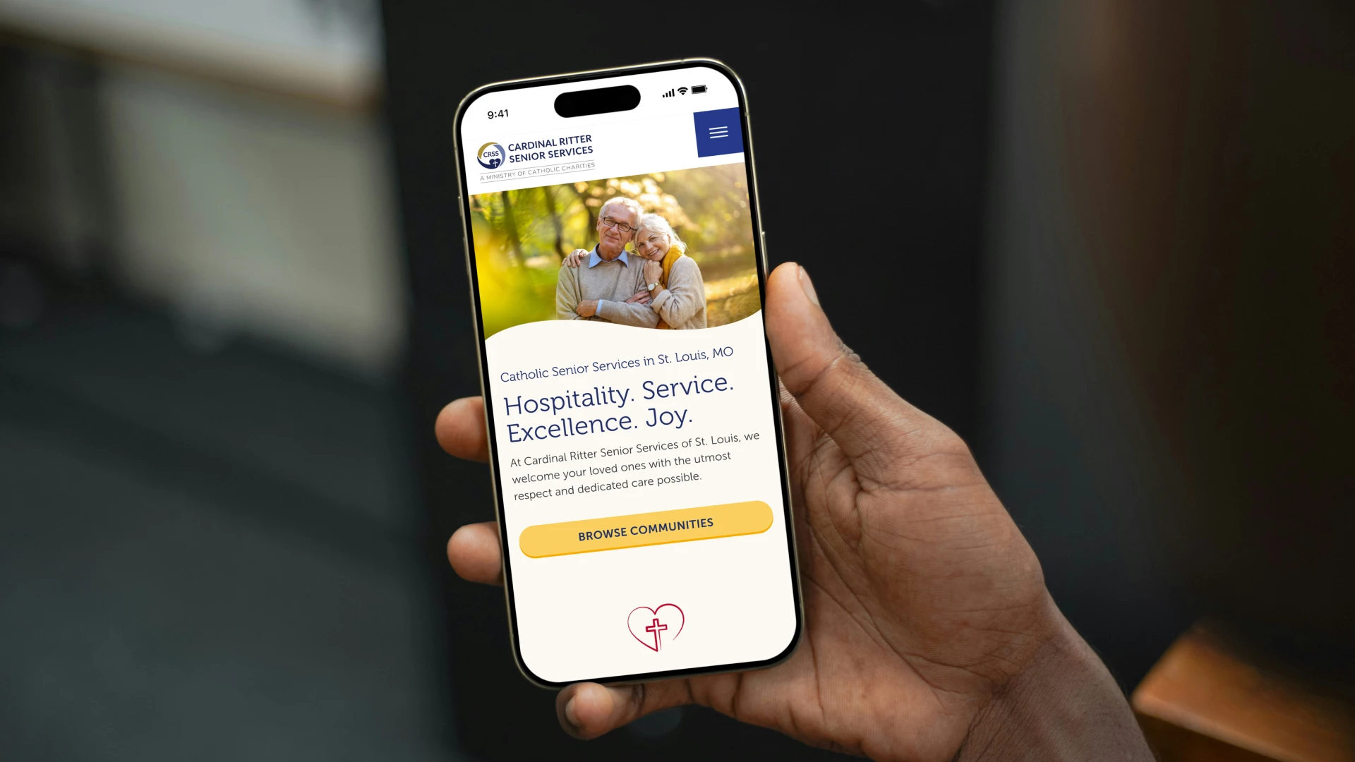 A person holds a smartphone displaying a Catholic senior services website. The screen shows an elderly couple smiling in a park, with text highlighting hospitality and service.