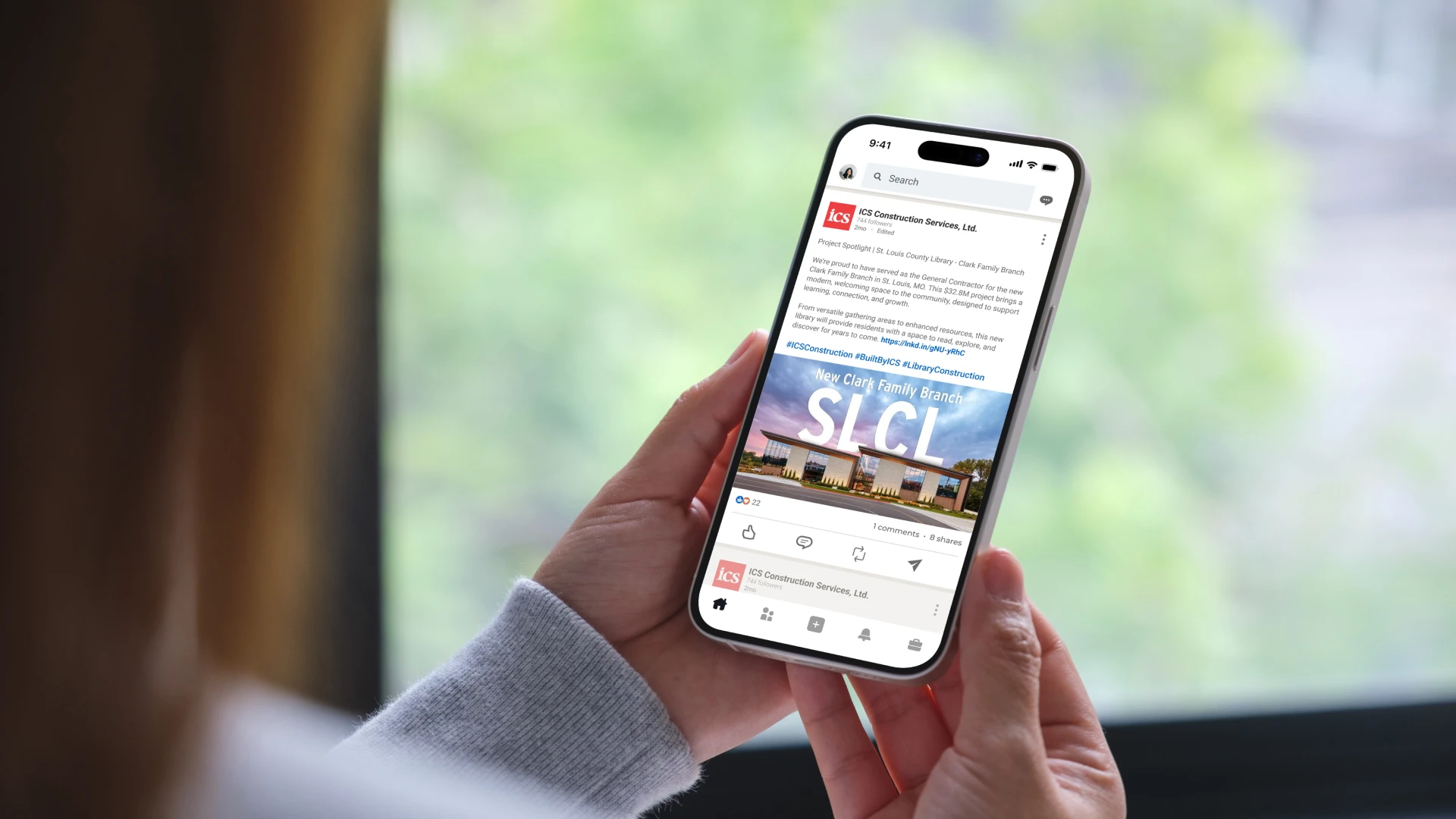 A person holding a smartphone displays a social media post featuring an office building and the text "SLCI." The background is softly blurred greenery.