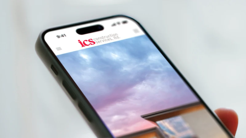 Smartphone held at an angle displaying the ICS Construction Services logo on screen with a photo of a building and colorful sky behind it.