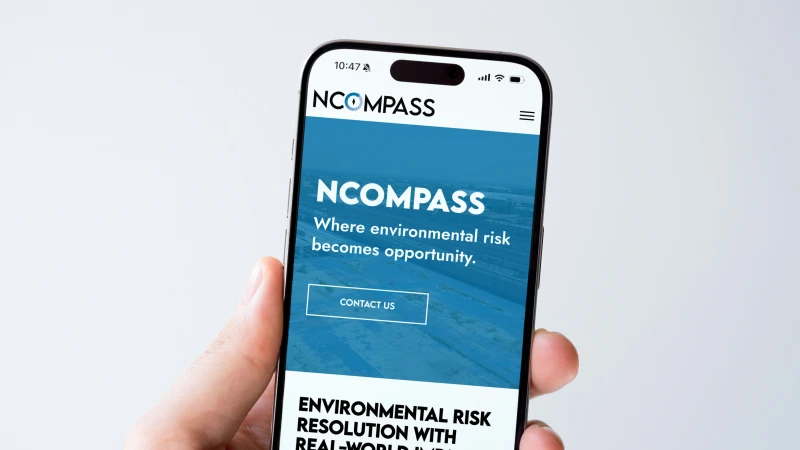 Smartphone screen showing the NCOMPASS website with the slogan 'Where environmental risk becomes opportunity' and a 'Contact us' button, held by a hand.