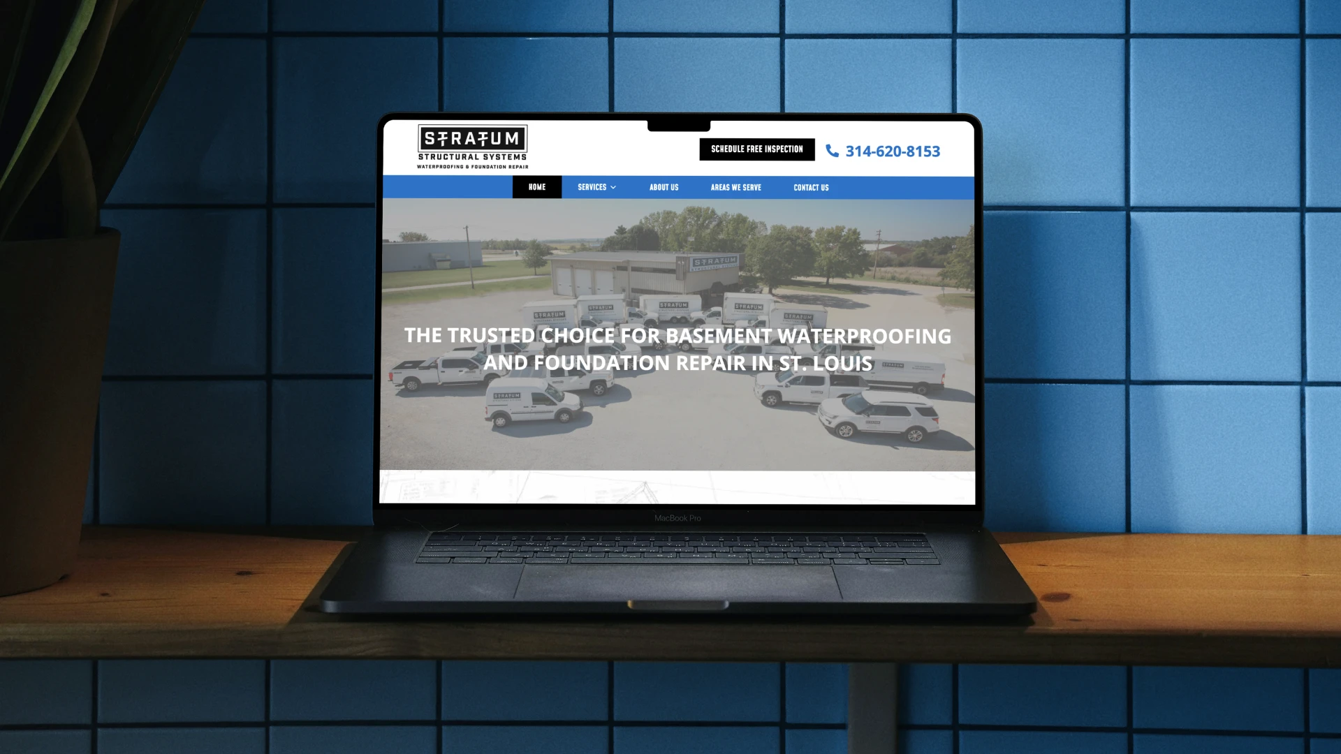 Front-facing shot of a black laptop sitting on a wooden desk against a blue tiled wall. The screen displays the Stratum Structural Systems homepage, featuring a large banner image of their service fleet and the headline, "The Trusted Choice for Basement Waterproofing and Foundation Repair in St. Louis."