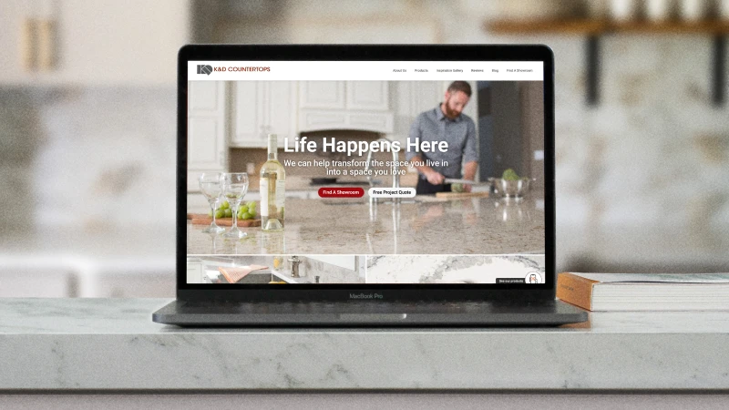 A laptop displays a website for K&D Countertops on a marble countertop. The site shows a kitchen scene with the text "Life Happens Here" conveying warmth.