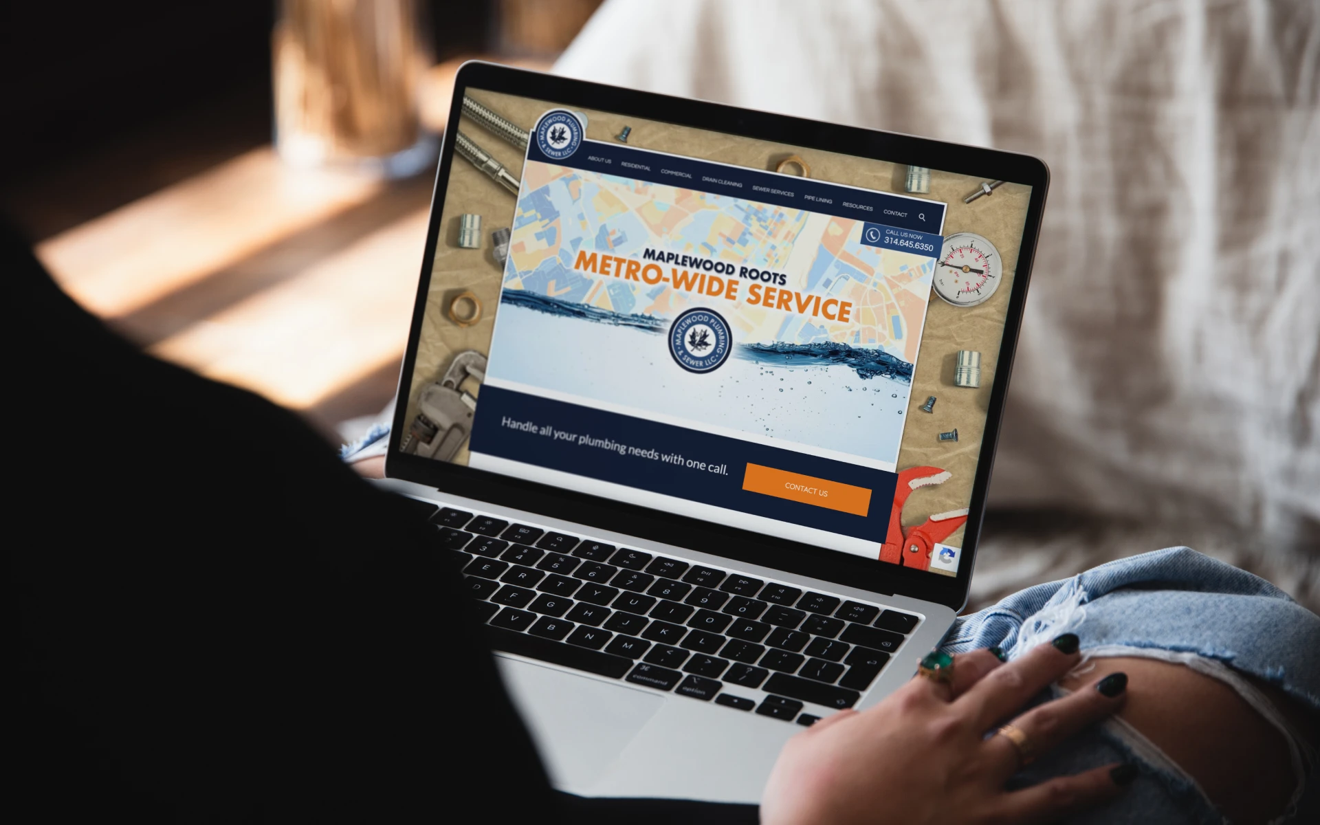 A person using a laptop displays a website for "Maplewood Roots Metro-Wide Service," featuring plumbing services. The scene is cozy and relaxed.