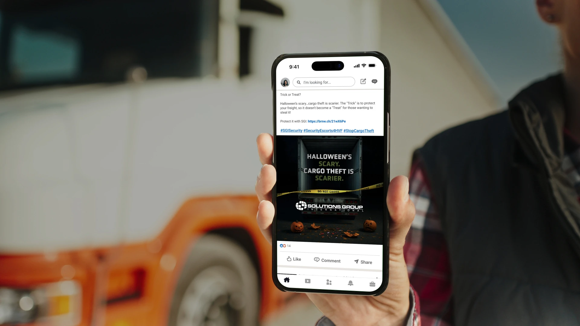 A person holds a phone showing a social media post with a Halloween theme. The text says "Halloween's scary. Cargo theft is scarier." A truck is blurred in the background.