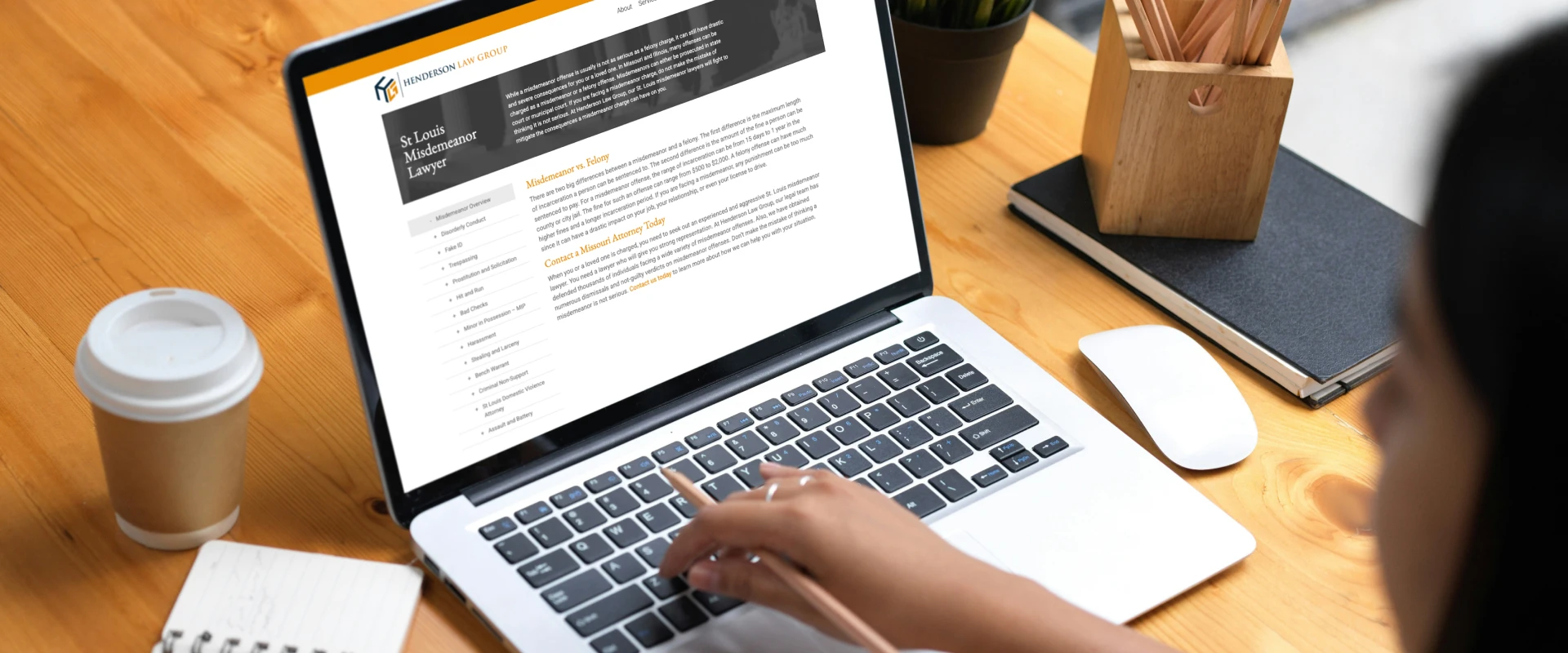 Person typing on a laptop displaying a law firm webpage about a St. Louis misdemeanor lawyer, on a wooden desk with a coffee cup nearby.